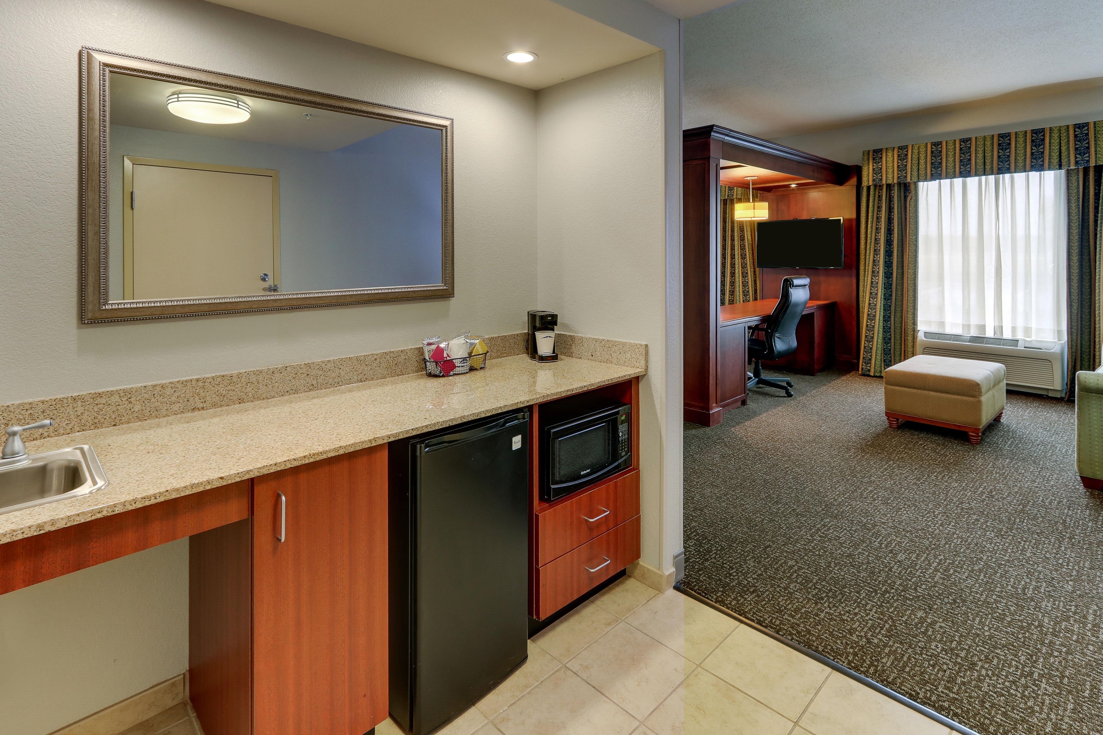 A hotel room with a kitchenette, a large mirror, a television, and a seating area with a sofa and a chair.