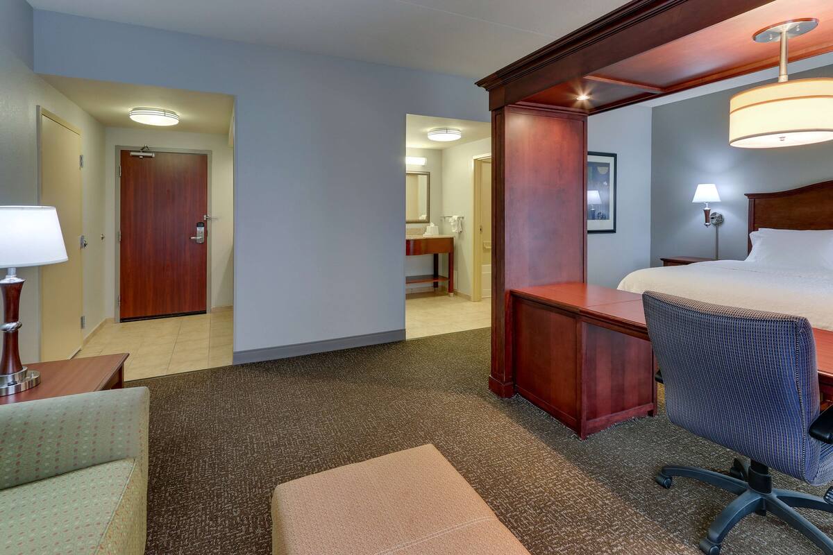 A hotel room with a bed, desk, chair, lamp, and a door leading to another room.