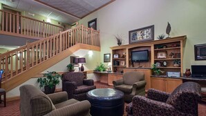 A spacious living area with a wooden staircase, comfortable seating, a flat-screen TV, and a bookshelf with various decorative items. - AmericInn by Wyndham Newton (Newton)