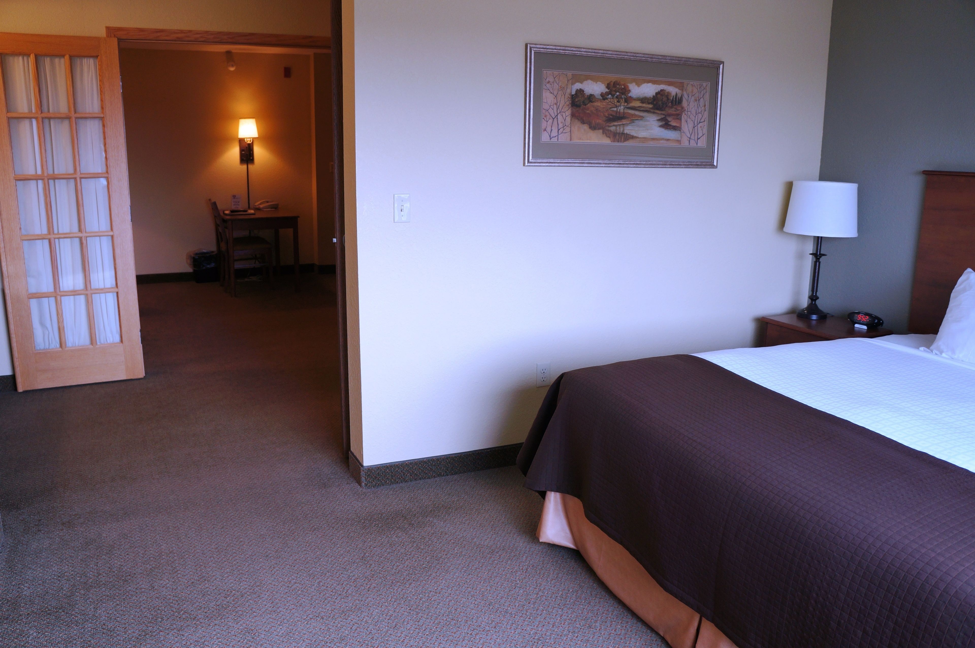 Habitación de hotel con cama, mesita de noche, lámpara, un cuadro en la pared y una puerta que da a otra habitación.