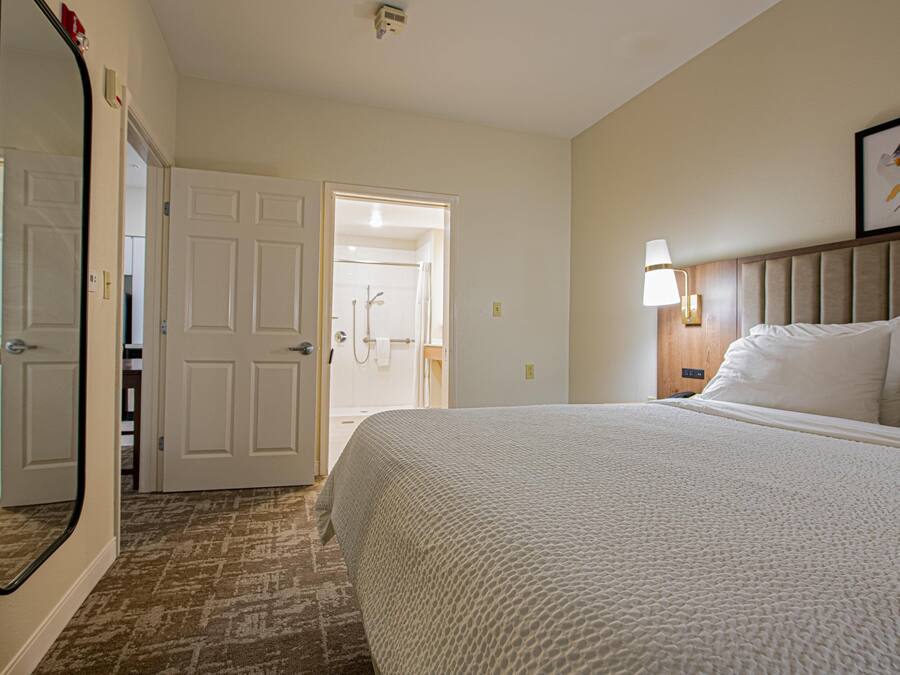 A hotel room with a large bed, a mirror, a door leading to a bathroom, and a nightstand with a lamp.