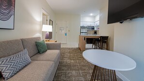 A compact living space with a sofa, a small table, a kitchen area with a microwave, and a TV mounted on the wall. - Staybridge Suites Lafayette by IHG (Lafayette)