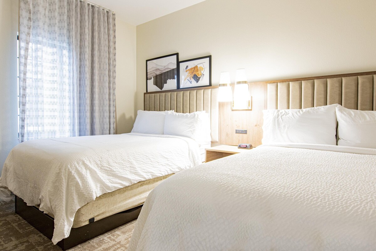 A hotel room with two beds, a headboard, a nightstand with a lamp, and two framed pictures on the wall.