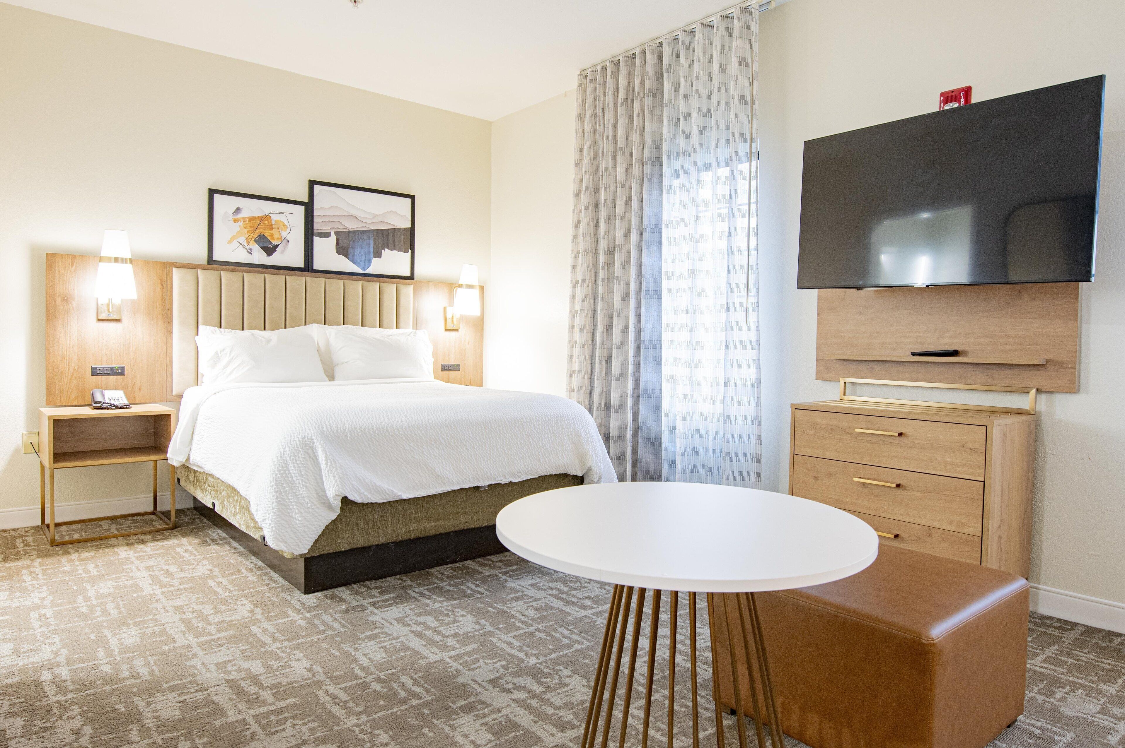 A hotel room with a bed, bedside tables, a TV, a small table, and a large window with curtains.