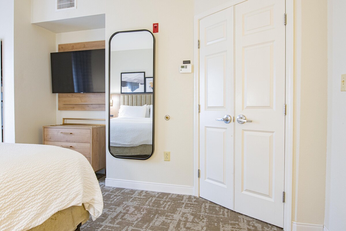 A hotel room with a bed, a dresser, a large mirror, a flat-screen TV, and a door.