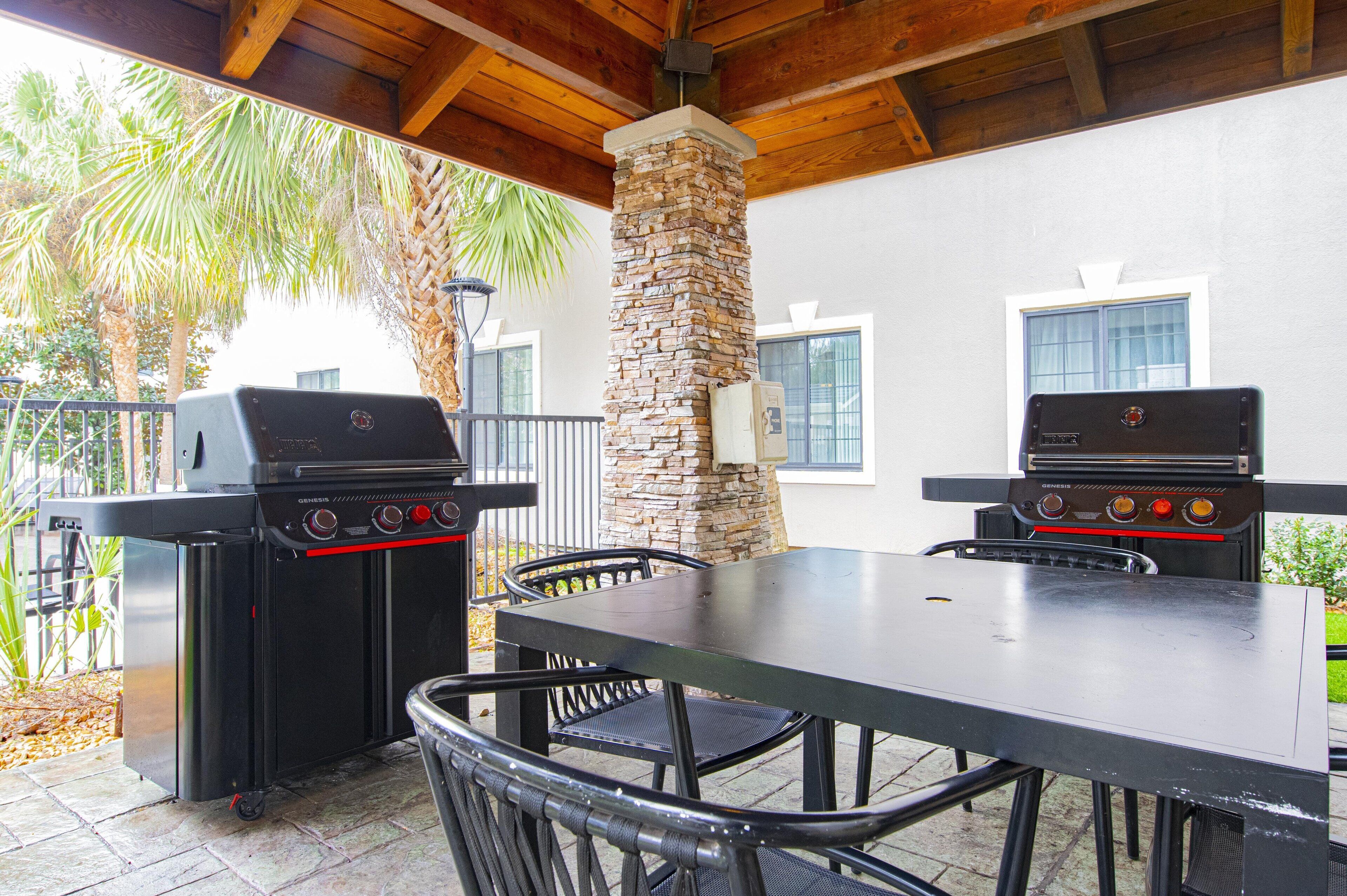 Een terras buiten met twee barbecues, een stenen pilaar en een tafel met stoelen.