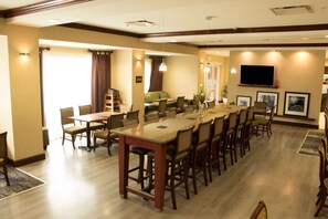 Un espace de restauration d’hôtel avec une longue table en bois, plusieurs chaises, un canapé et un téléviseur fixé au mur.