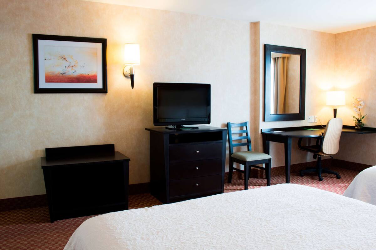 Habitación de hotel con cama, escritorio, silla, televisor, cómoda, espejo y lámpara de pared.