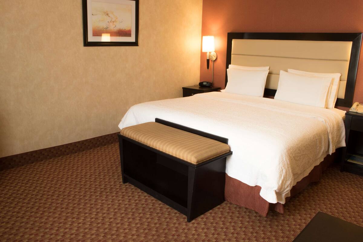 Una habitación de hotel con una cama grande, una mesita de noche, un banco y una lámpara fijada a la pared.