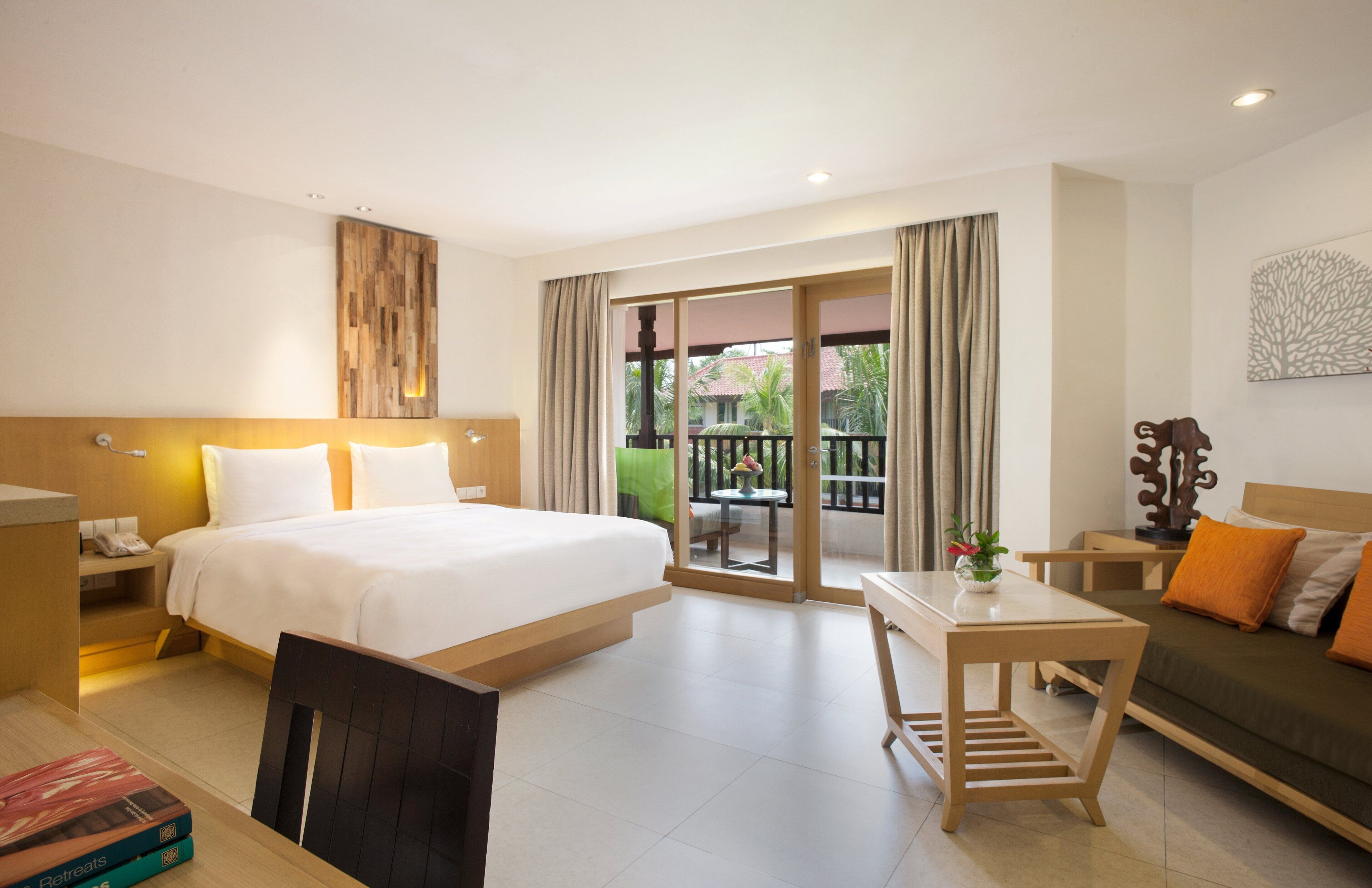 A hotel room with a bed, a desk, a sofa, a balcony with a view, and a wooden headboard.