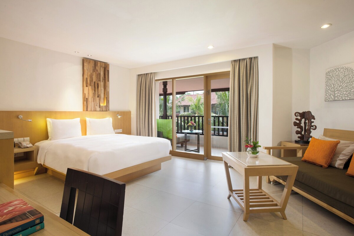 A hotel room with a bed, a desk, a sofa, a balcony with a view, and a wooden headboard.