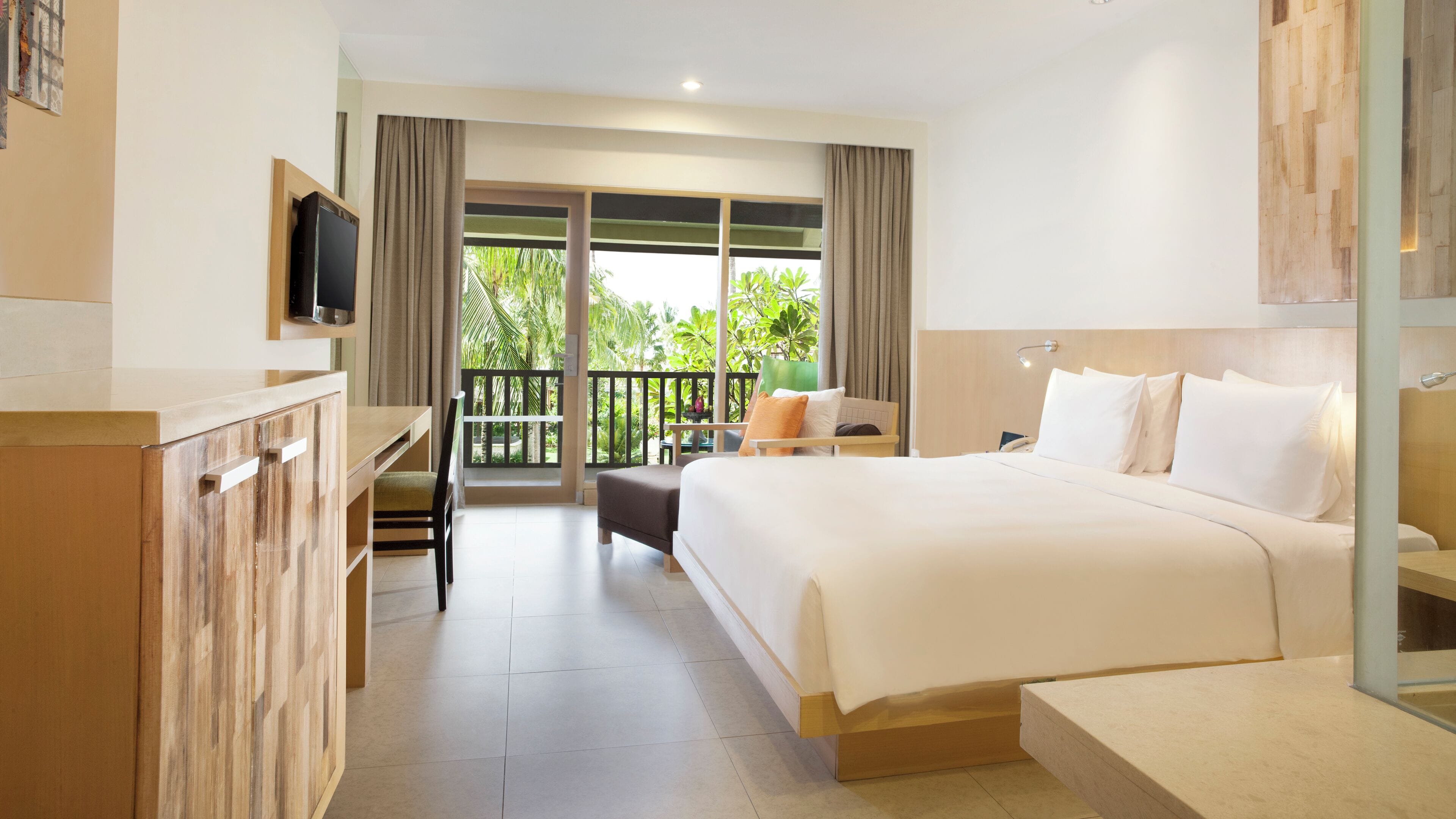 A hotel room with a bed, a desk, a TV, and a balcony with a view of greenery.