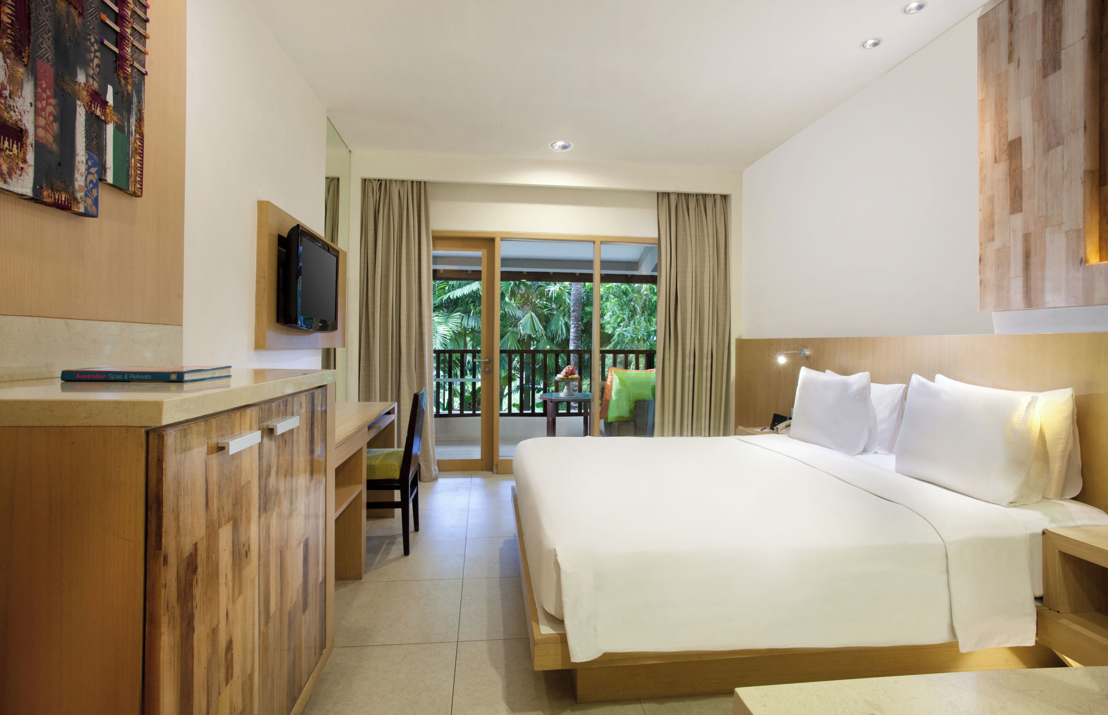 A hotel room with a large bed, a desk with a television, and a balcony with a view of greenery.