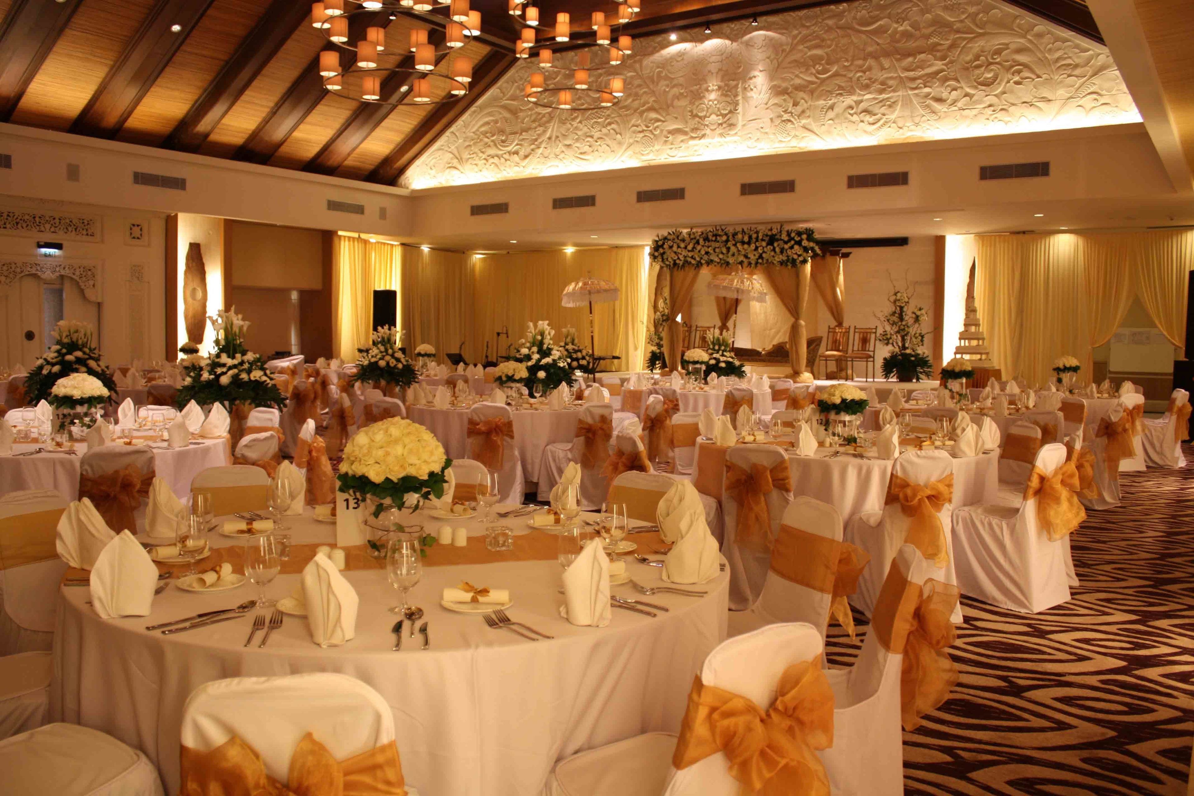 A banquet hall with round tables set for a formal event, featuring white tablecloths, gold sashes, and floral centerpieces.