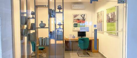 A reception area with a desk, computer, and a green chair. There are glass partitions with decorative items and a sign indicating the business center.