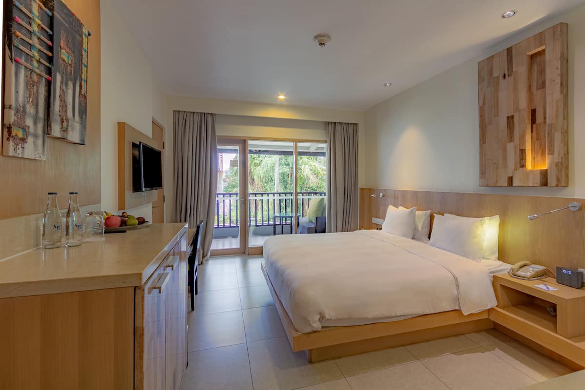 A modern hotel room with a large bed, a desk with a flat-screen TV, a balcony with curtains, and a wooden headboard.