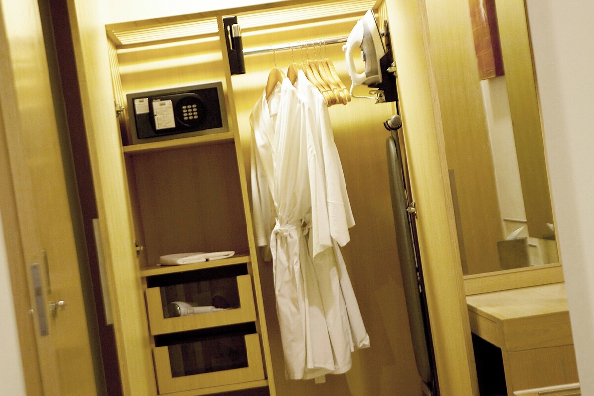 A hotel room with a wardrobe containing hanging clothes, a microwave, and a shelf with a tray.