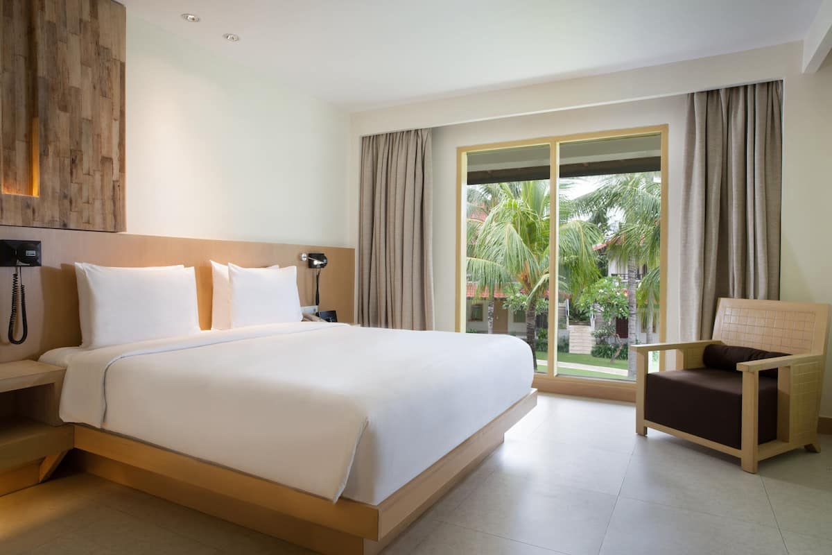 A hotel room with a large bed, a bedside table with a phone, a chair, and a view of palm trees through the window.