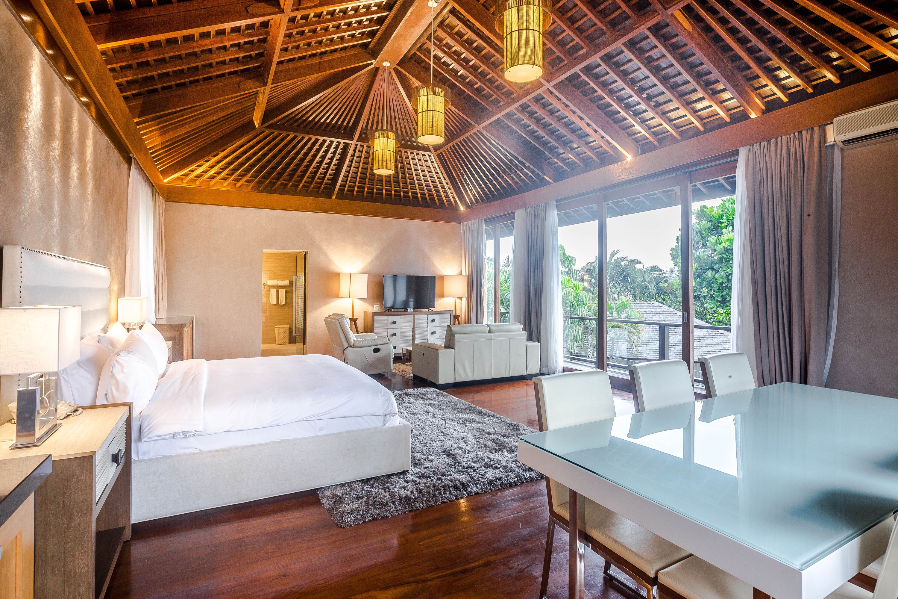 A hotel room with a large bed, a desk, a chair, a TV, and a balcony with a view of greenery.