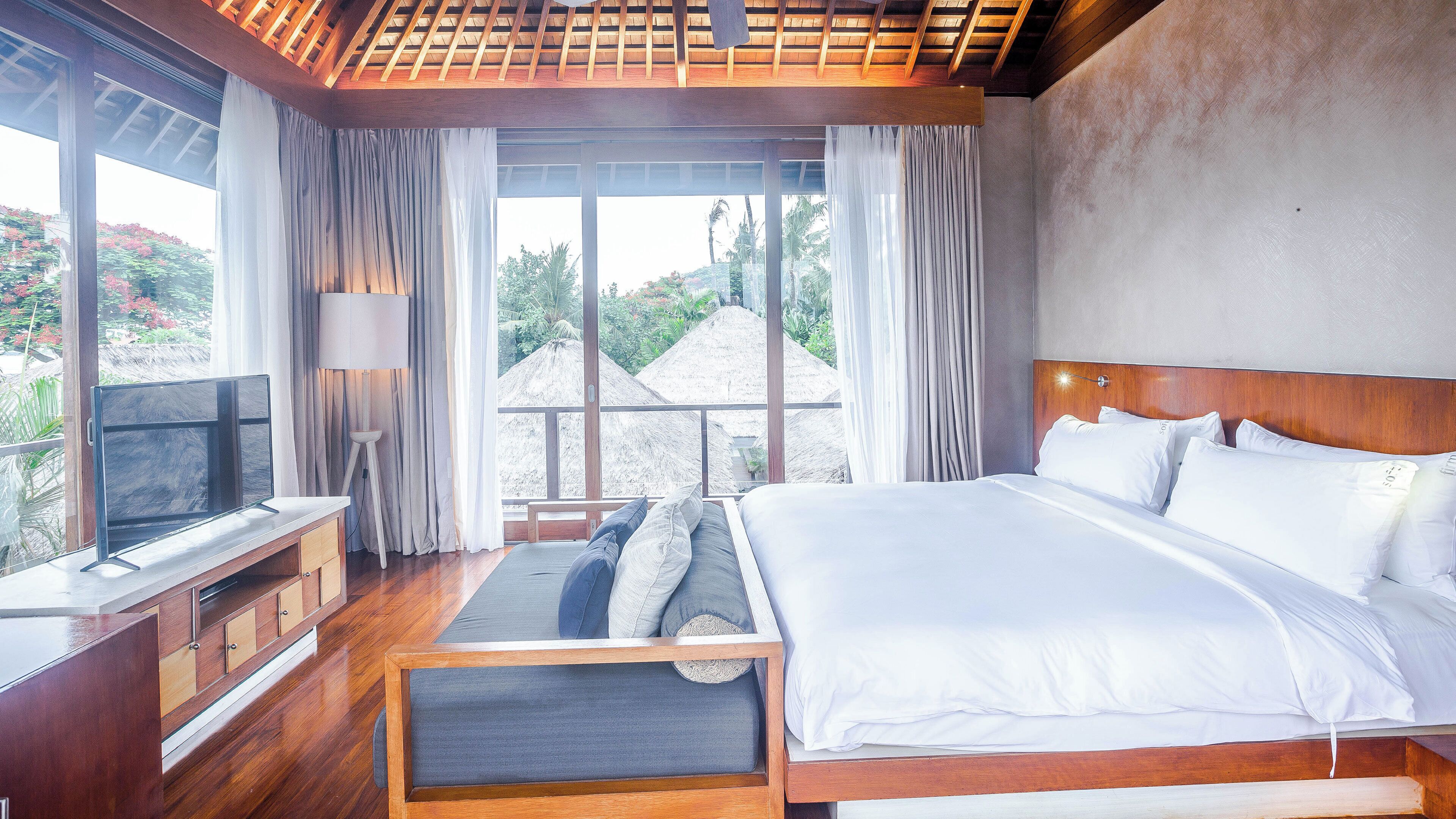 Quarto de hotel moderno com uma cama grande, televisão e vista para uma paisagem tropical.