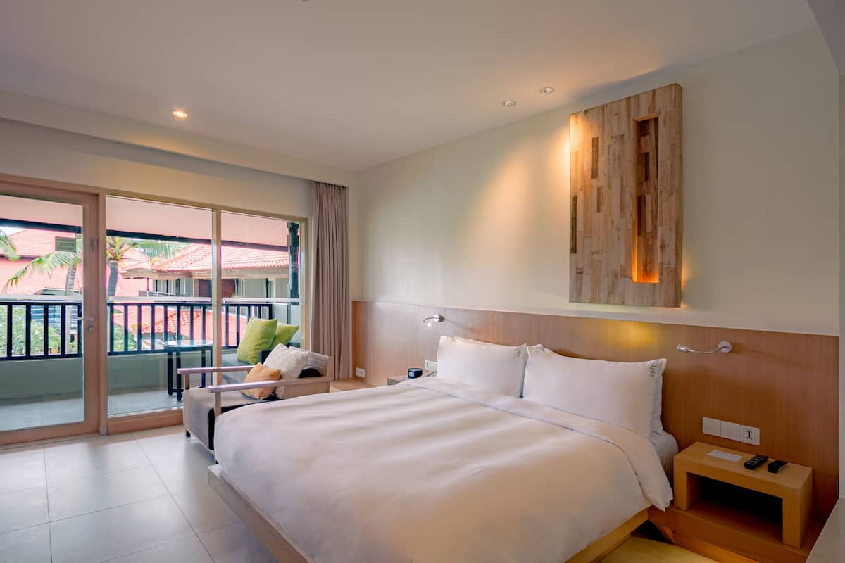 A hotel room with a large bed, a wooden headboard, a nightstand, a desk, and a balcony with a view of palm trees and a building.