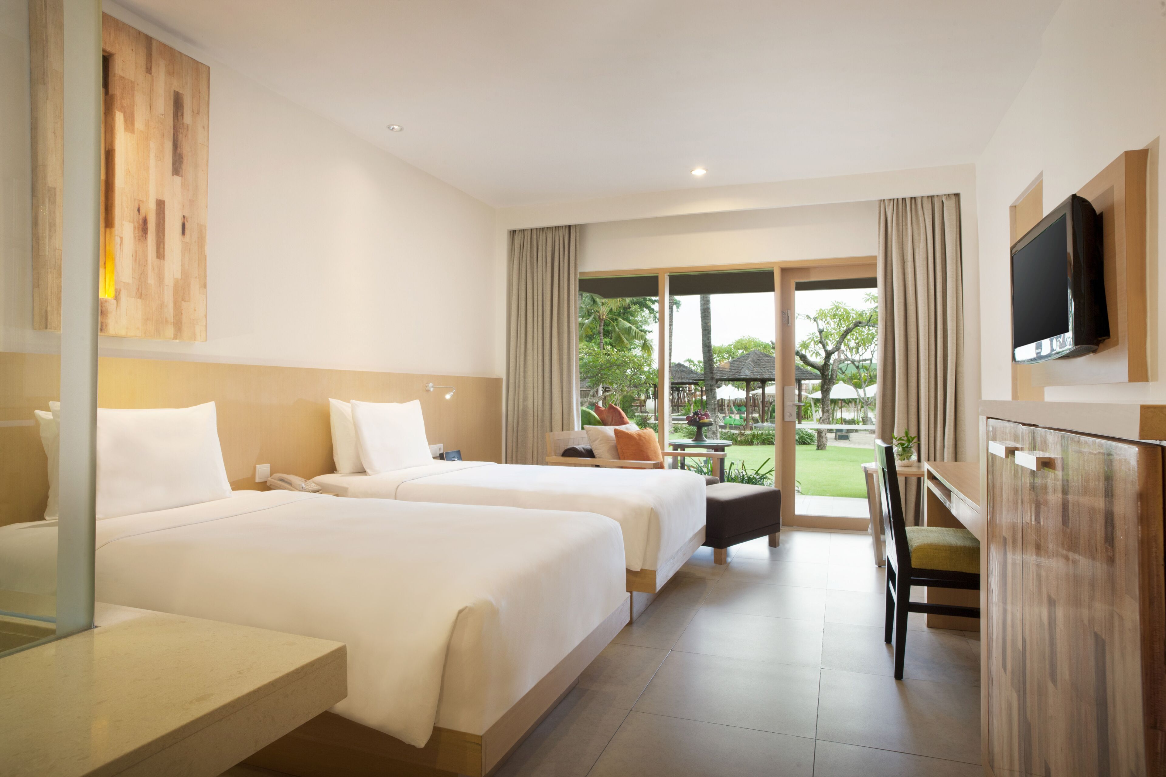 A hotel room with two beds, a desk, a chair, a TV, and a view of the garden.