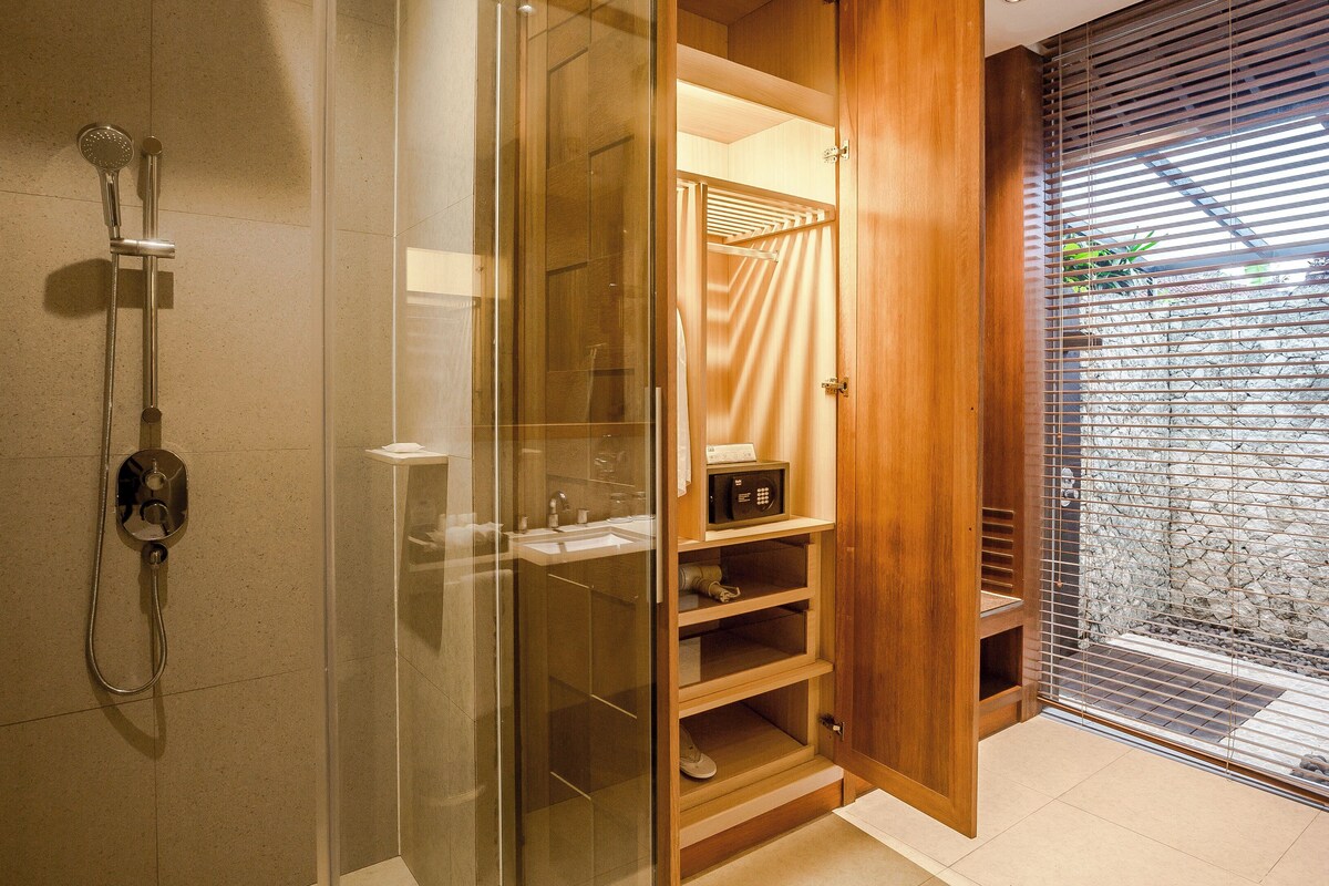 A modern bathroom with a glass-enclosed shower, wooden shelving, and a view of a stone wall through a window with blinds.
