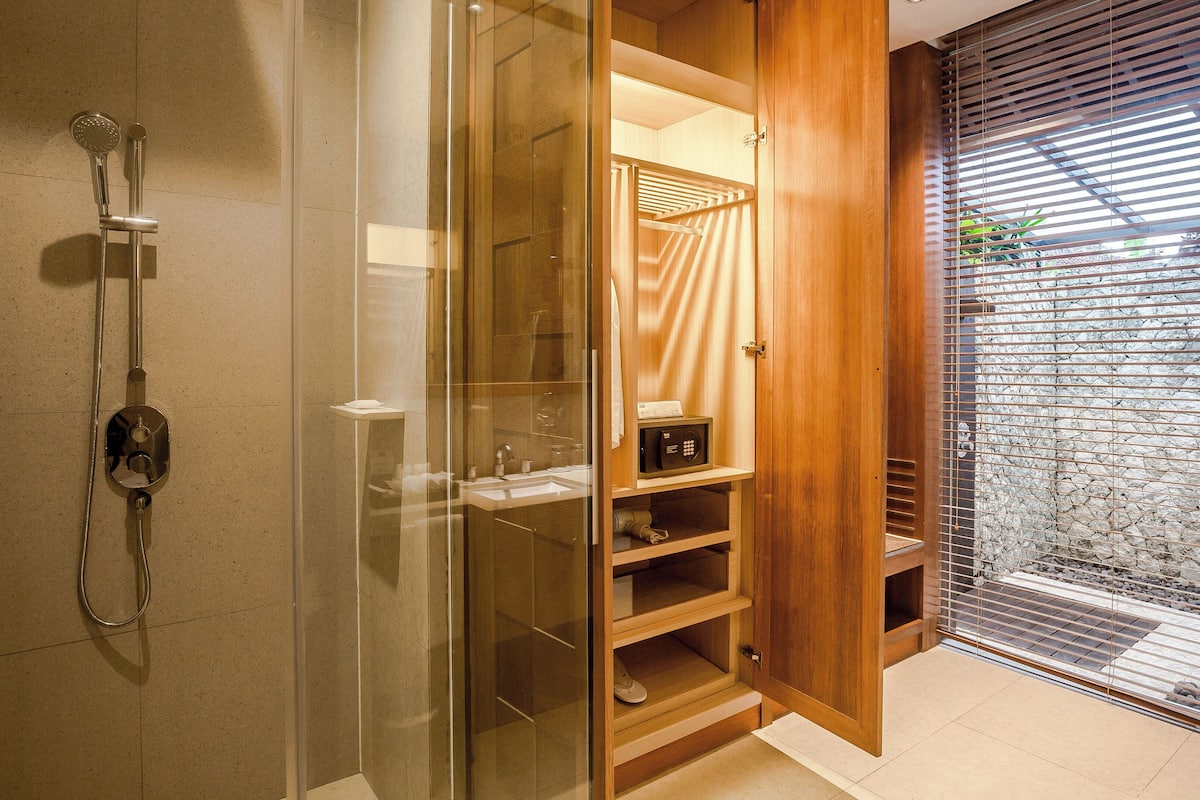 A modern bathroom with a glass-enclosed shower, wooden shelving, and a view of a stone wall through a window with blinds.