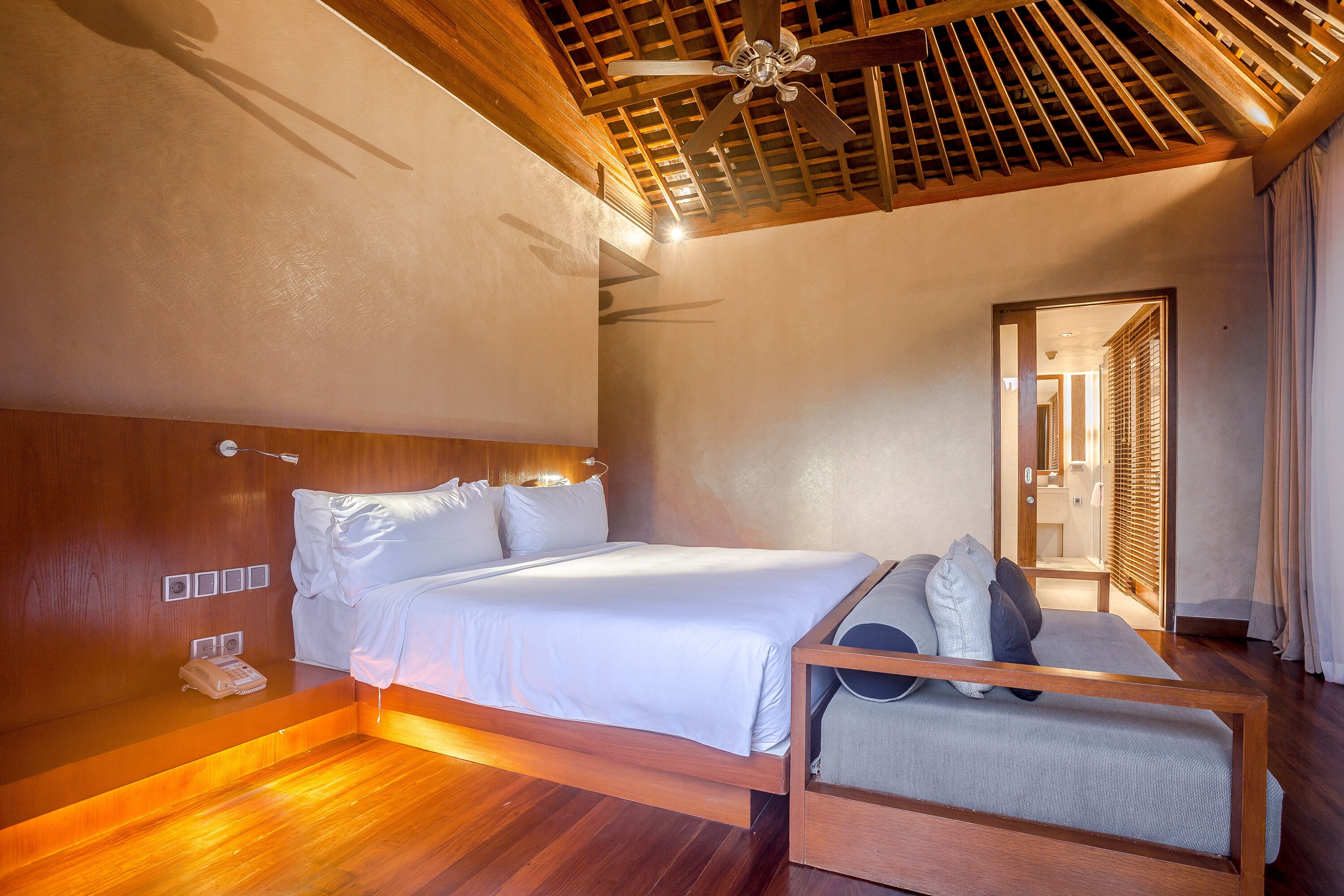 Une chambre d’hôtel avec un plafond en bois, un lit recouvert de draps blancs, un banc en bois agrémenté de coussins et un plancher en bois.