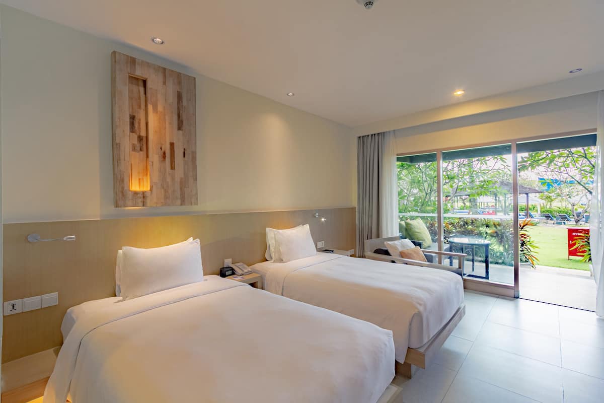 A hotel room with two beds, a wooden accent wall, a large window with a view of the outdoors, and a balcony with a table and chairs.