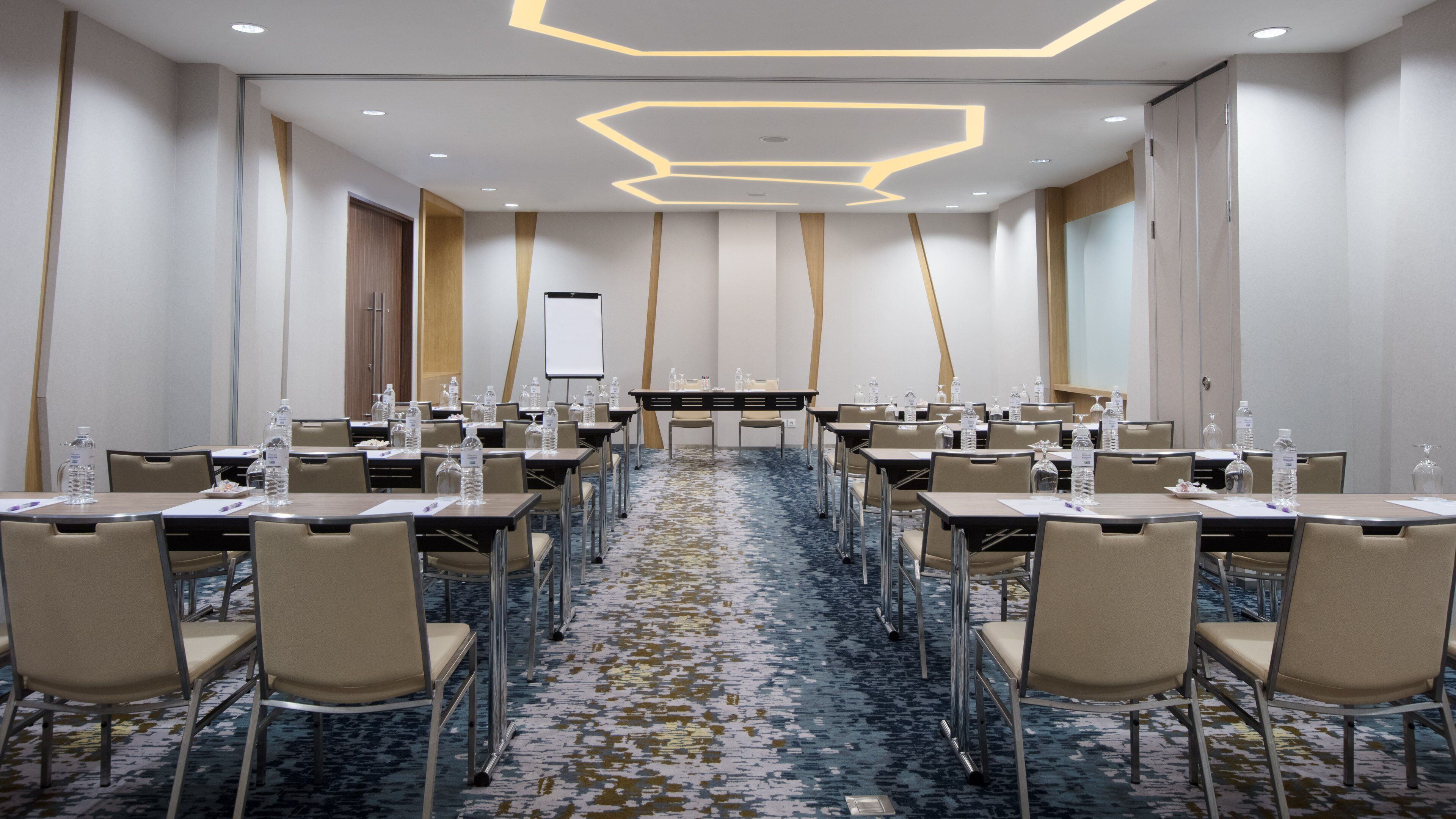 Une salle de conférence équipée de tables, de chaises, de bouteilles d’eau et d’un écran de présentation.