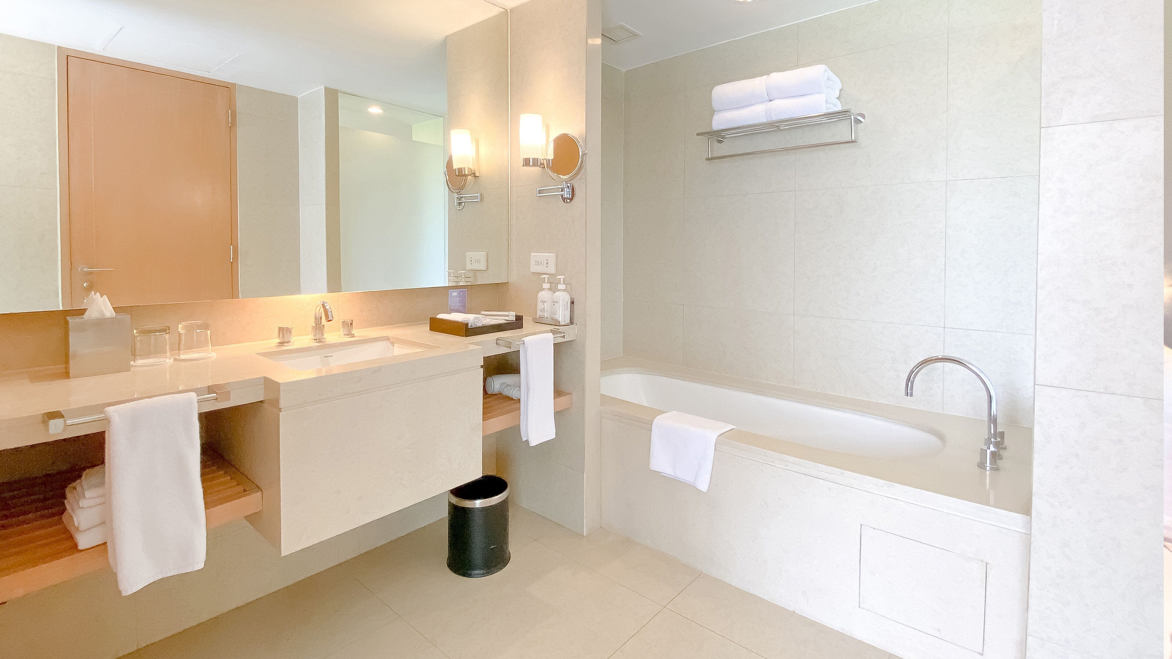 A modern bathroom with two sinks, a large mirror, a bathtub, and a trash can.
