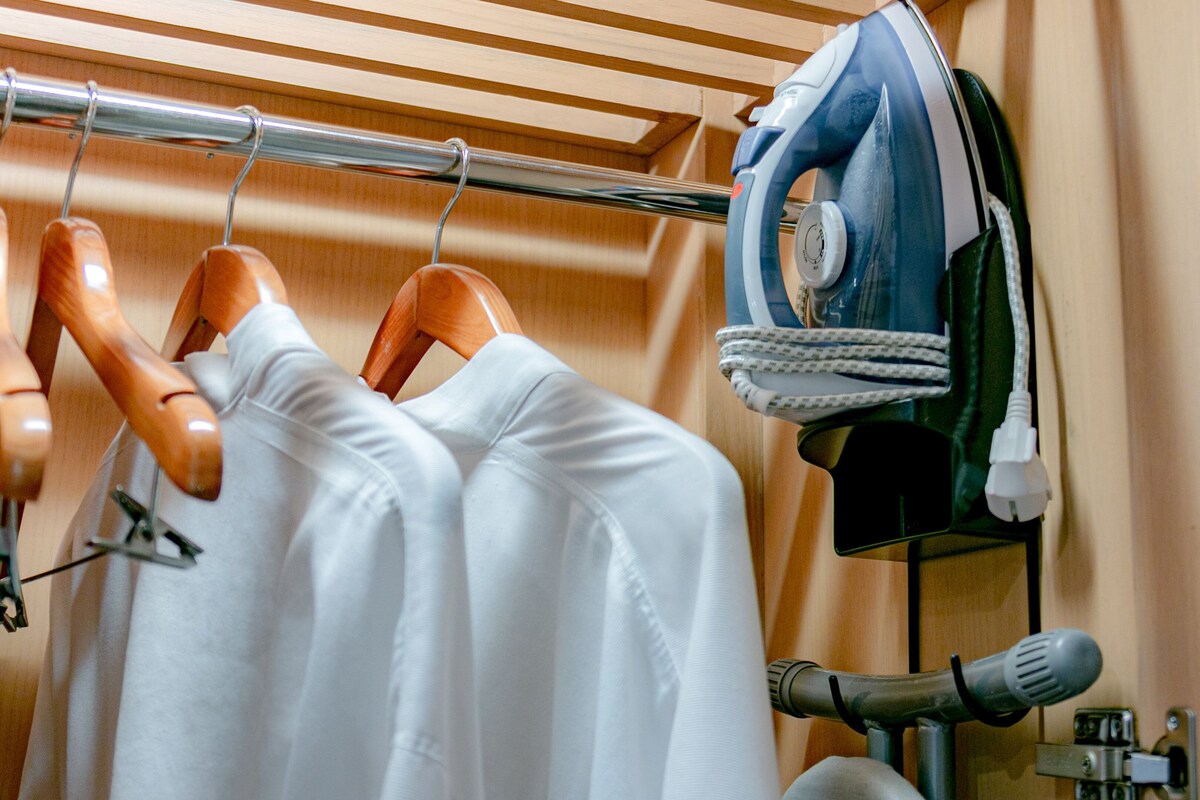 A clothing rack with hangers and a pair of jeans, an ironing board with an iron, and a wooden chair.