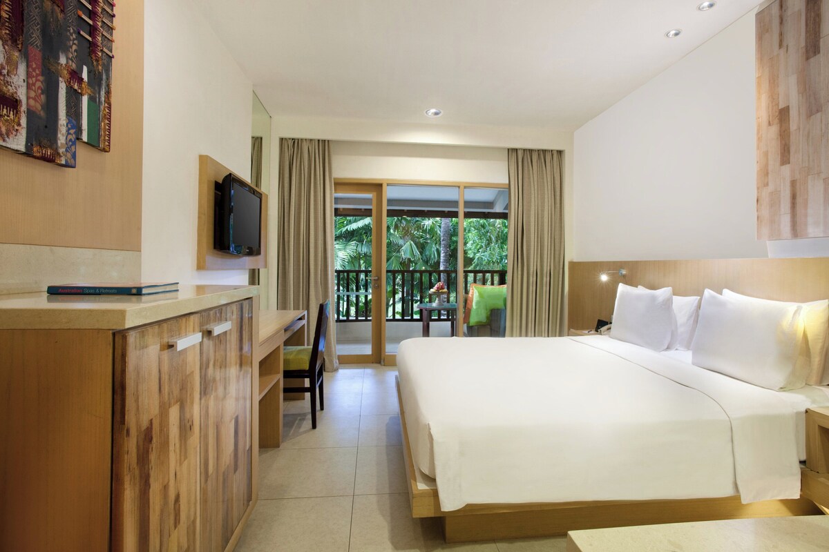 A hotel room with a bed, a desk, a television, and a balcony with a view of greenery.