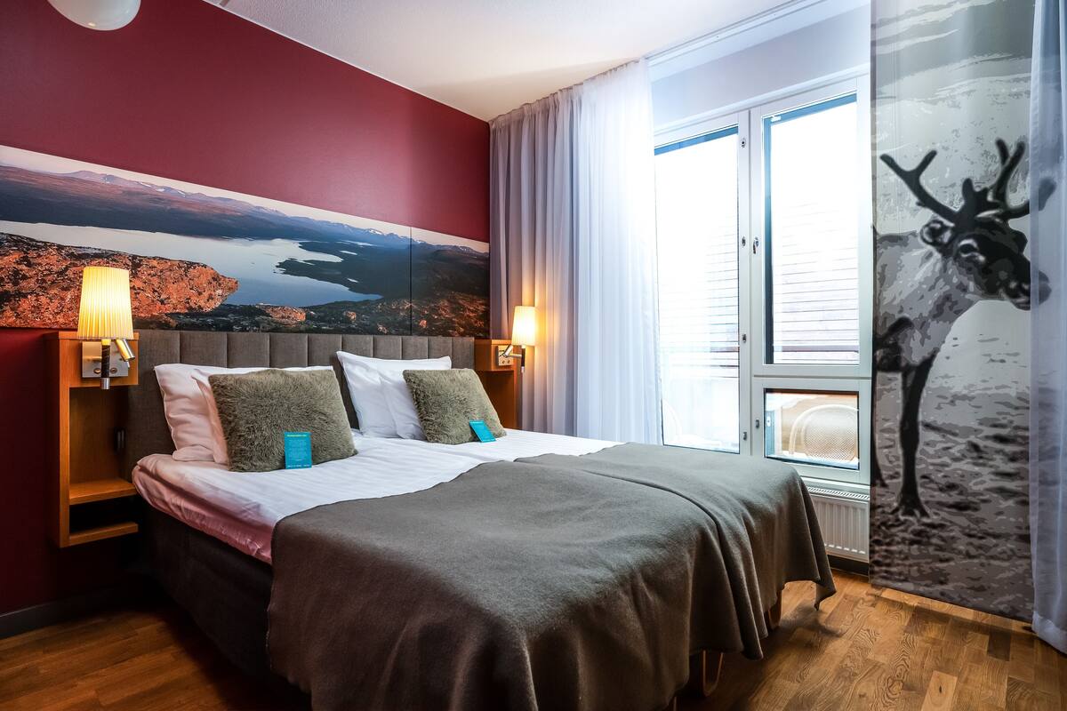 A hotel room with a bed, two bedside tables, a nightstand, a large window with curtains, and a wall painting.