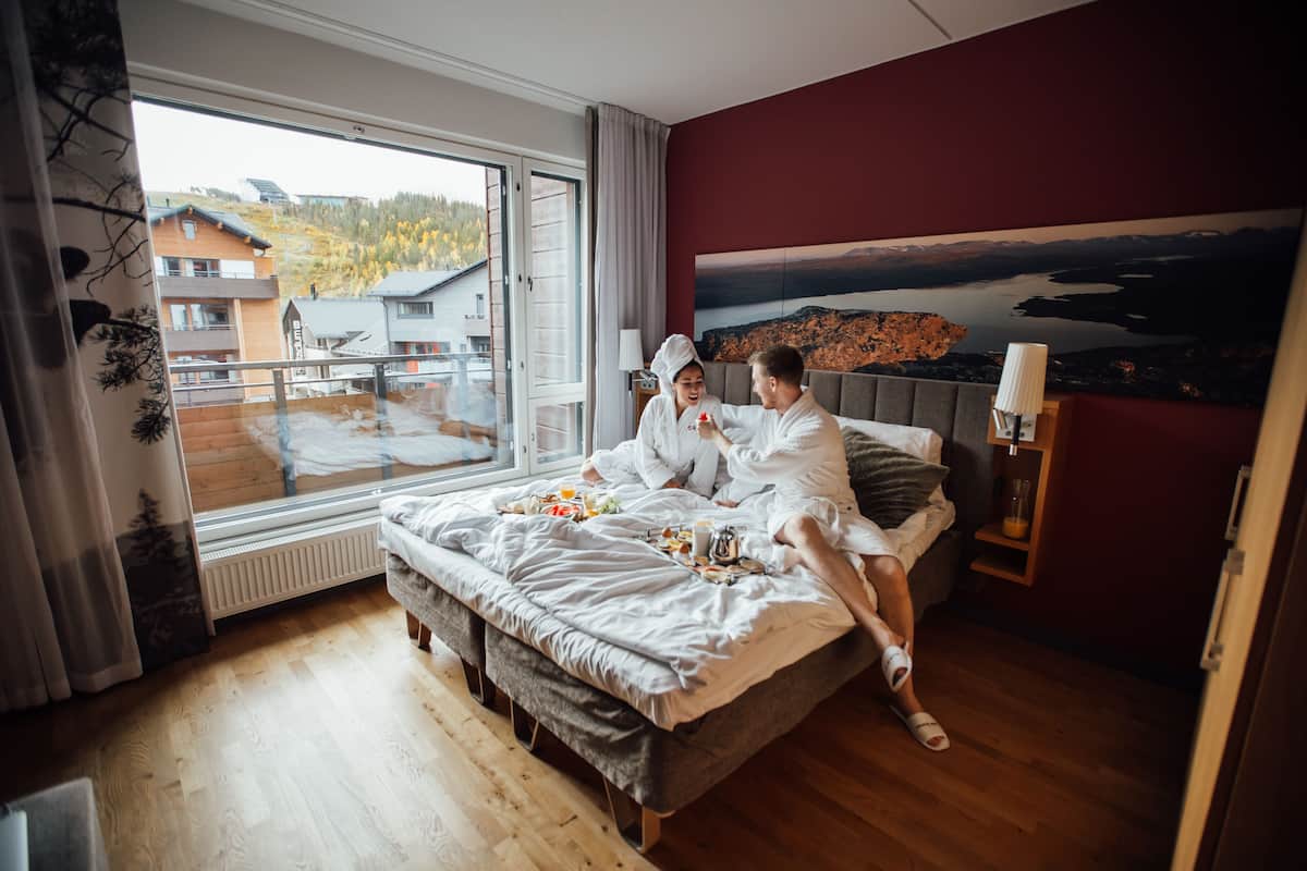 A bedroom with a large window, a bed, a nightstand, and a painting on the wall.