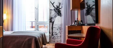 A hotel room with a bed, a red chair, a wooden table, a wall-mounted TV, and a large window with curtains.