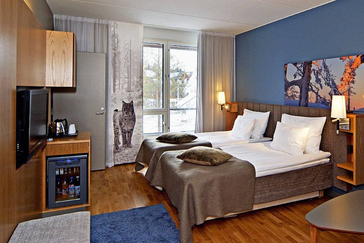 A hotel room with a bed, a TV, a minibar, and a painting on the wall.