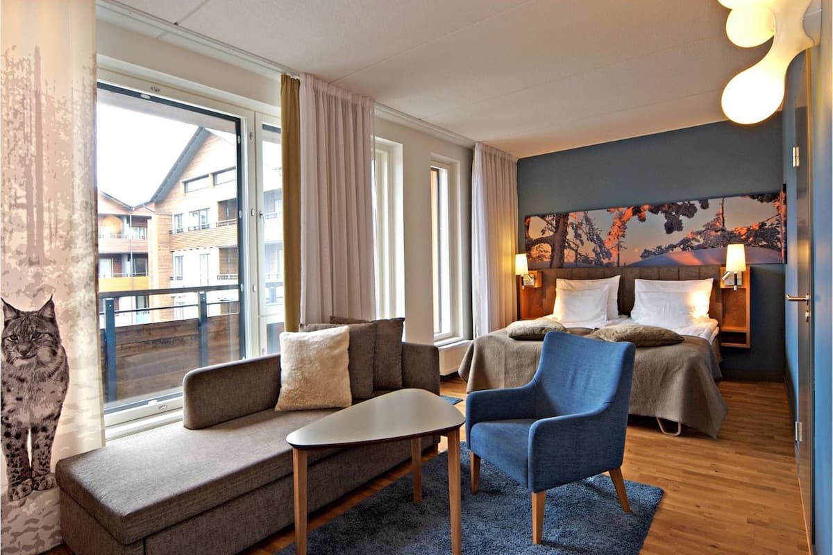 A modern hotel room with a sofa, a chair, a small table, and a bed with a headboard. There is a window with curtains, a wall with a painting, and a lamp on the bedside table.