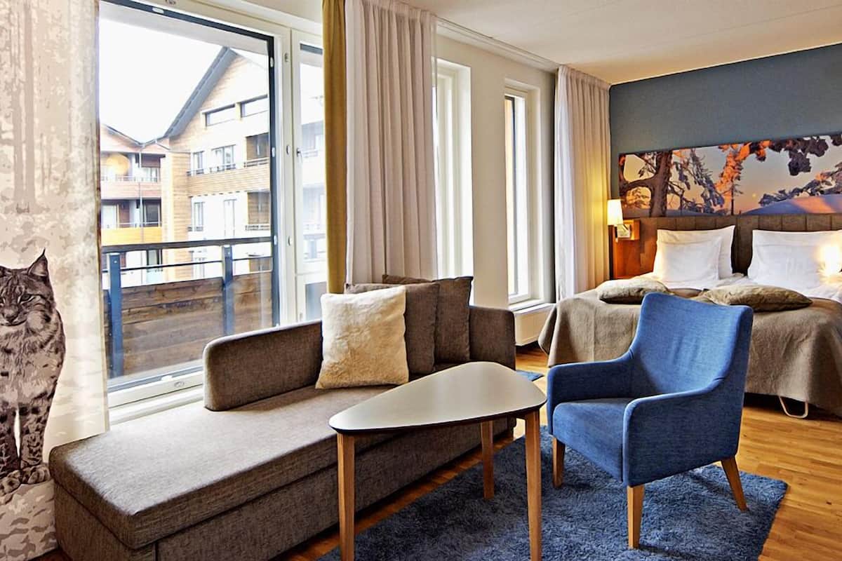 A hotel room with a sofa, armchair, and a bed with two pillows. There is a window with curtains, a painting on the wall, and a lamp on the nightstand.