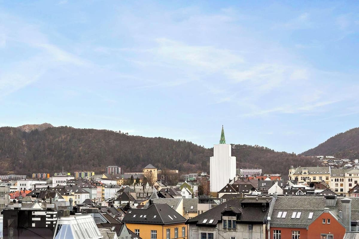 Un paisaje urbano con una torre blanca destacada, rodeada de varios edificios y montañas al fondo.