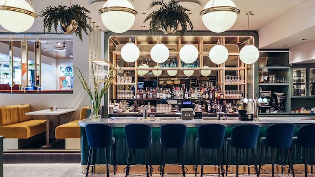 Una zona de bar con una barra alargada, varios taburetes y lámparas colgantes.