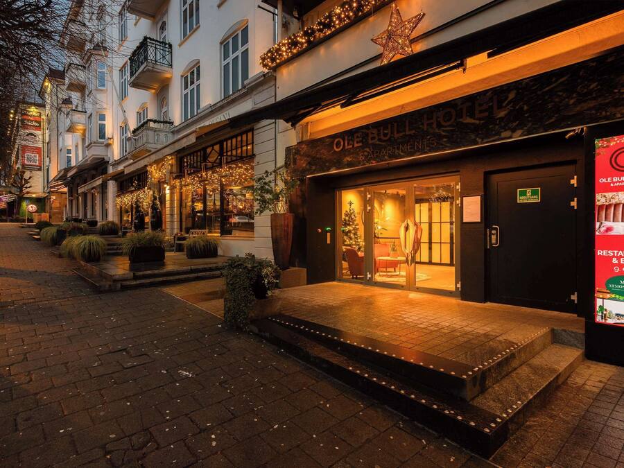 A hotel entrance with a sign for 'Ole Bull Hotel & Apartments,' a brick pathway, and outdoor seating.