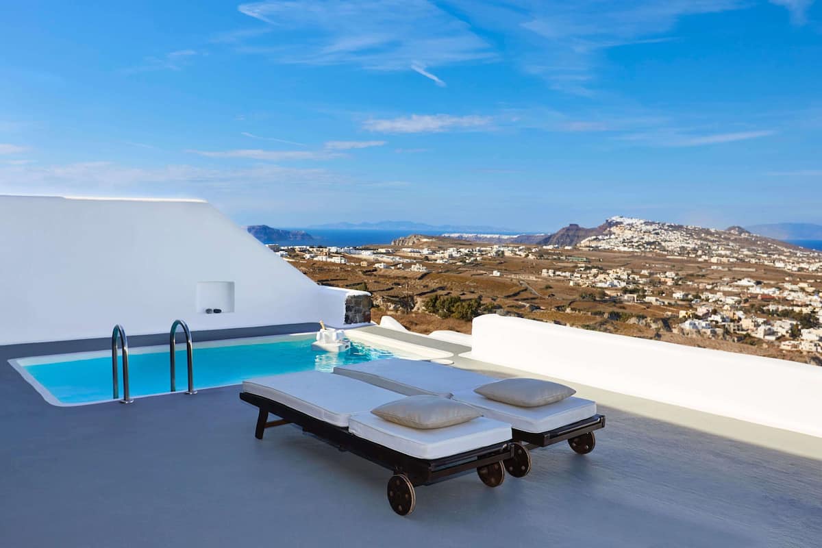 A poolside area with a view of a coastal town, a lounge chair, and a small pool.