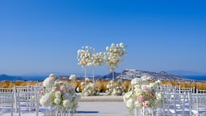Un lugar para bodas al aire libre con sillas blancas y arreglos florales, con un cielo azul despejado y montañas al fondo.