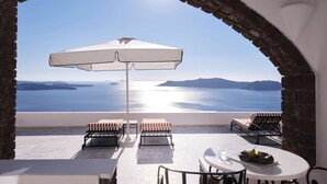 A terrace with a white umbrella, two lounge chairs, a table with a bowl of fruit, and a view of the sea and islands. - San Antonio (Santorini)