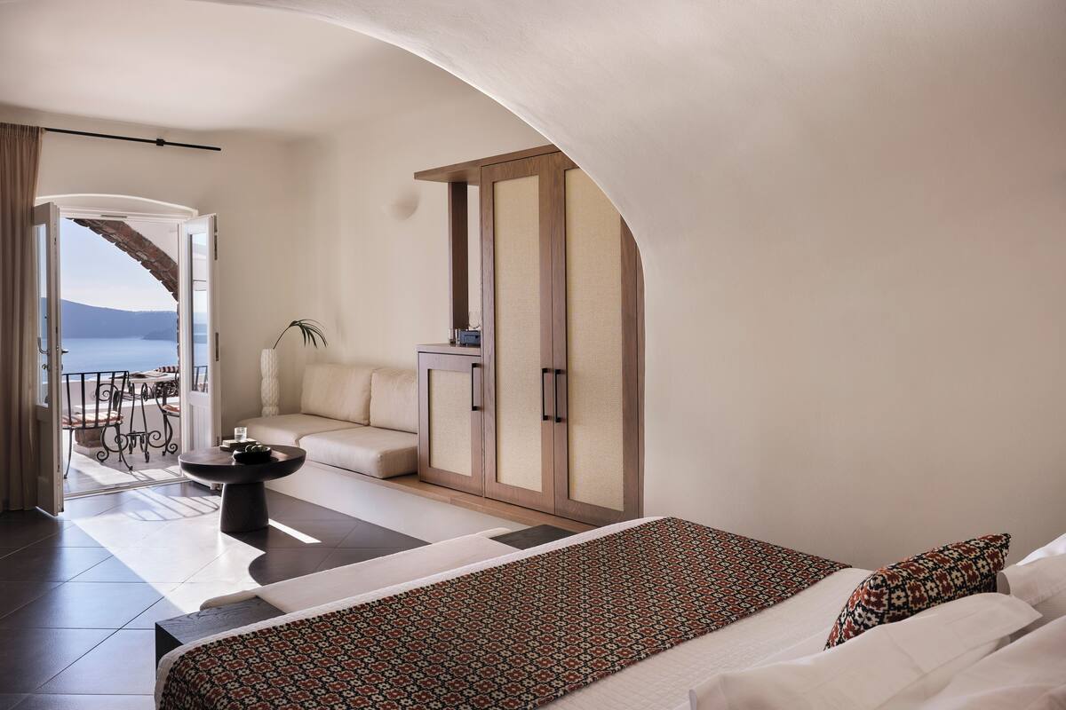Una habitación de hotel moderna con una cama grande, un área de descanso con sofá y sillones, una mesita y vista al mar a través de una puerta abierta.