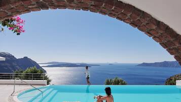 A pool with a clear view of the sea and mountains, a person standing by the pool, and a person sitting on a lounge chair.