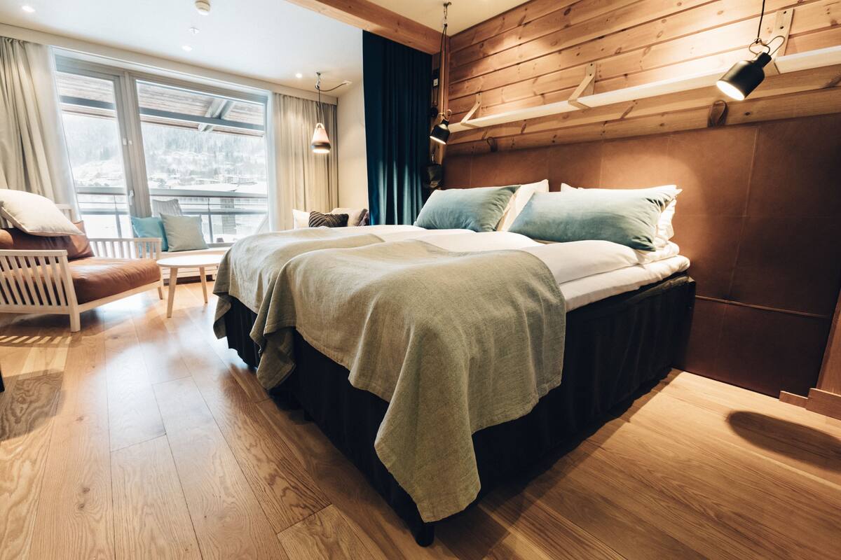 A modern bedroom with a large bed, wooden headboard, and a view of a snowy landscape through large windows.