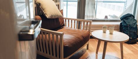 A wooden chair with a cushion, a small round table with two cups, and a backpack near a window with curtains.
